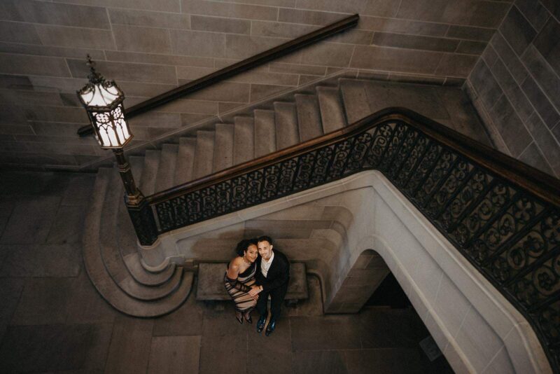 Elegant couple sitting on stairs at high-end venue with ornate railings and chandelier, showcasing luxury wedding decor and sophisticated ambiance.