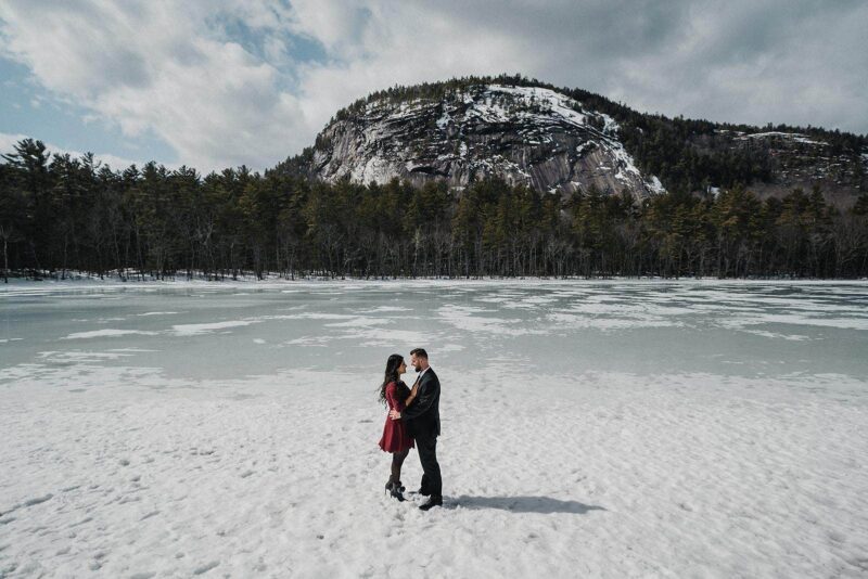Snow-covered lake with romantic couple in formal attire, surrounded by winter landscape and mountain backdrop, perfect for luxury winter wedding photos.