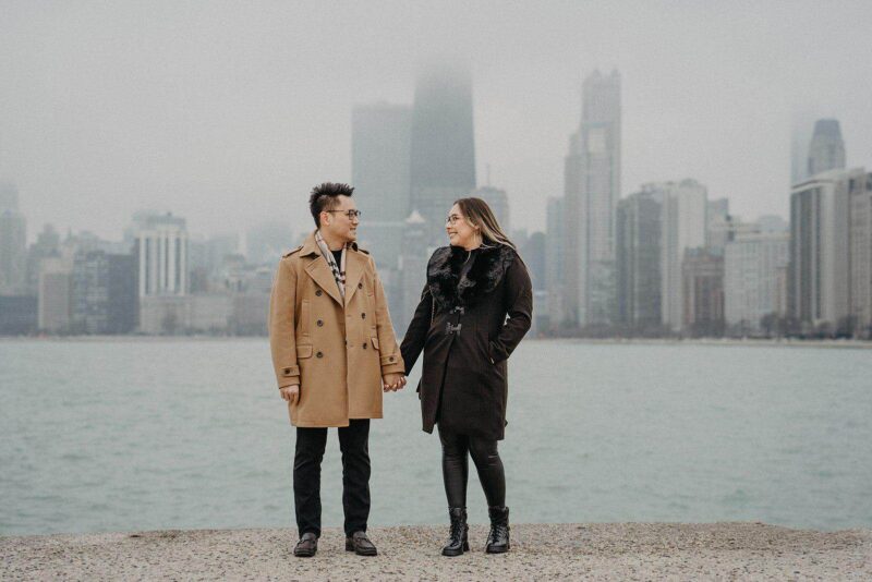 Elegant couple holding hands by the waterfront with a city skyline in the background.