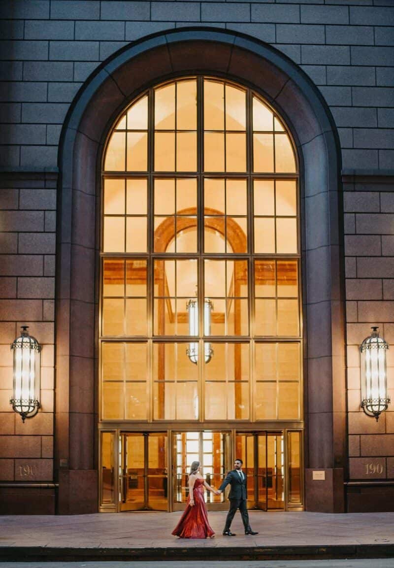 Elegant couple holding hands outside a luxurious venue with a grand glass entrance at dusk.