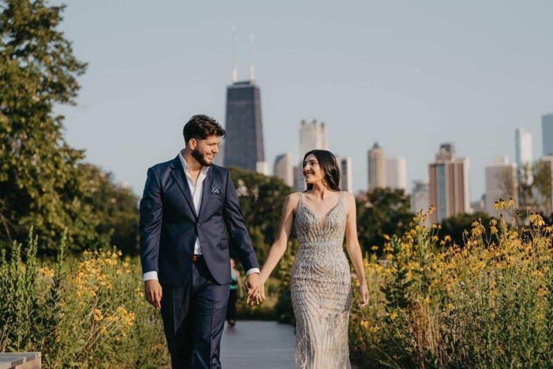 Elegant couple walking hand-in-hand in a luxurious urban park with Chicago skyline backdrop.