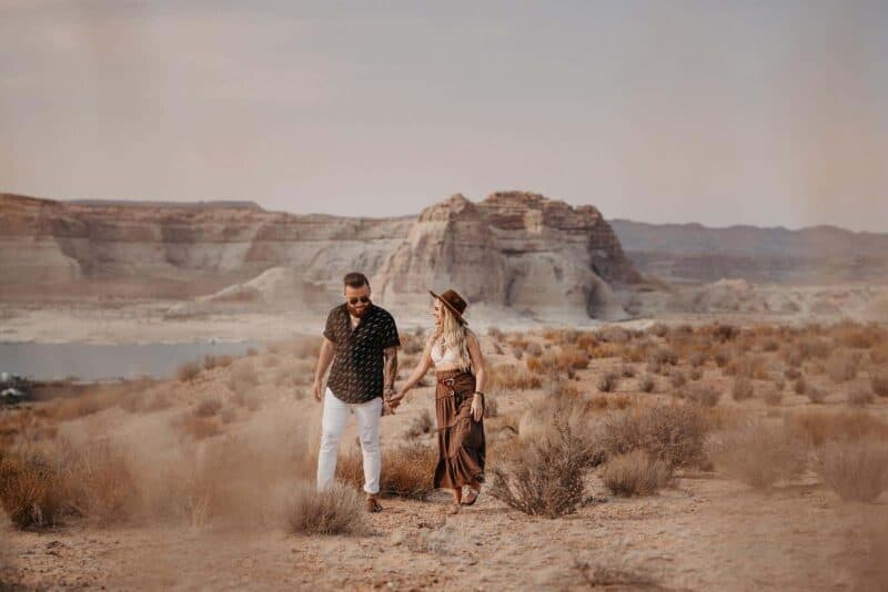Romantic couple walking hand in hand through a desert landscape with dramatic rock formations and a serene lake, perfect for high-end wedding inspiration.