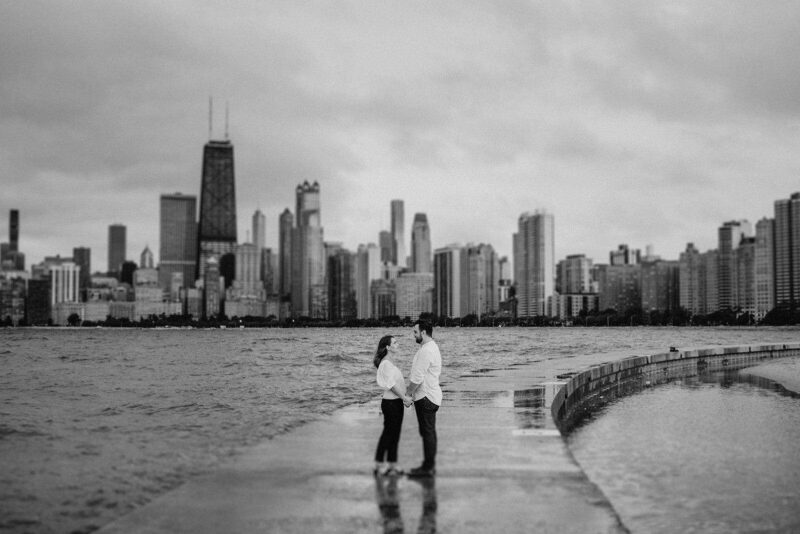 City skyline with tall skyscrapers and romantic couple holding hands on lakeside promenade in black and white image.