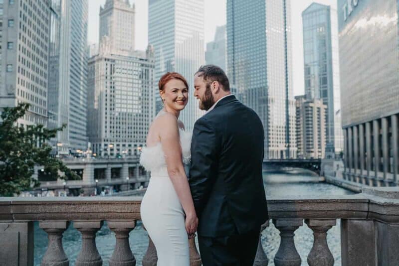 Elegant couple in wedding attire on city balcony with skyscrapers in the background.