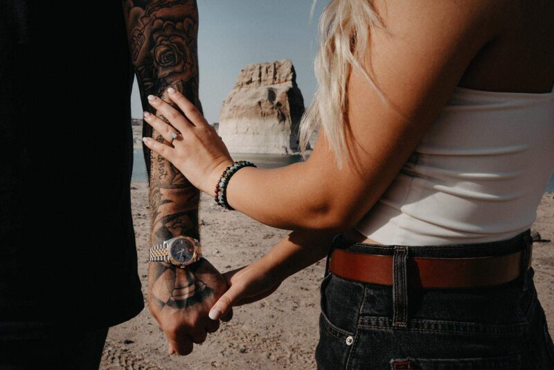 Engagement ring on woman's hand, couple holding hands at the beach, with rock formation in background.
