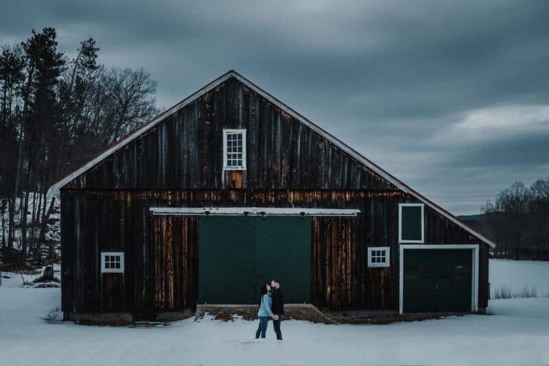 Rustic barn wedding venue in winter with couple kissing outside at sunset, high-end wedding location, elegant outdoor celebration, winter wedding decor, luxury rustic venue.