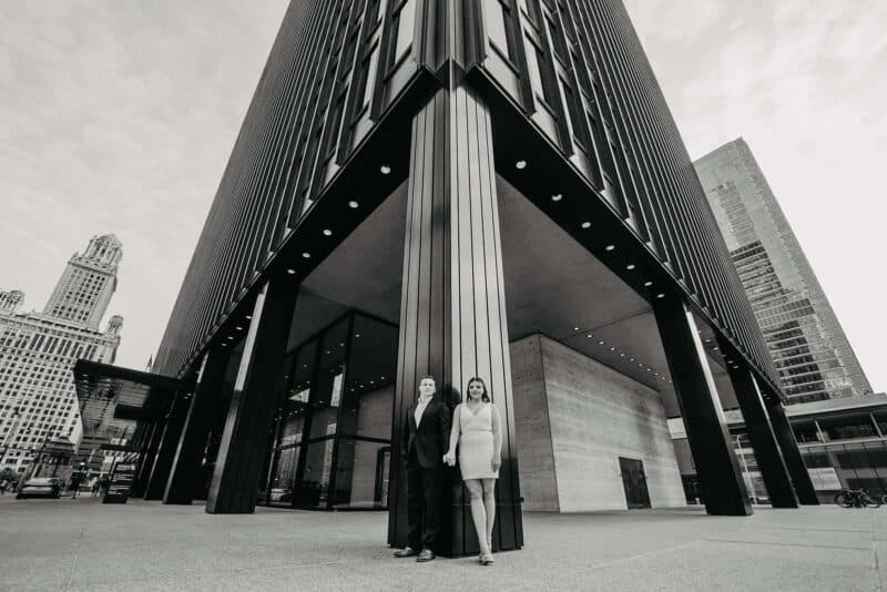 Elegant couple in formal attire standing outside a sleek, modern high-rise building in a luxurious cityscape setting.
