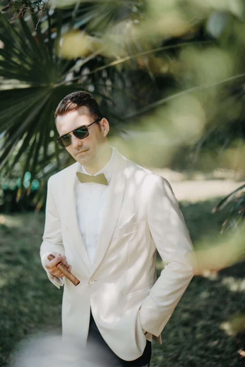 Elegant groom in a white tuxedo with a black bow tie and sunglasses, holding a cigar, outdoors amidst lush greenery at a luxury private estate.