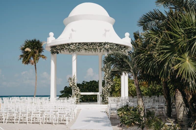 Chic outdoor wedding setup on a white sandy beach with a decorative white gazebo surrounded by lush tropical palm trees under a clear blue sky.