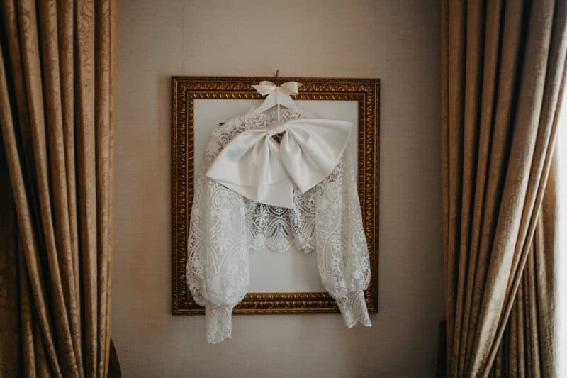 Elegant white lace wedding dress with a large satin bow hanging on a decorative gold frame.