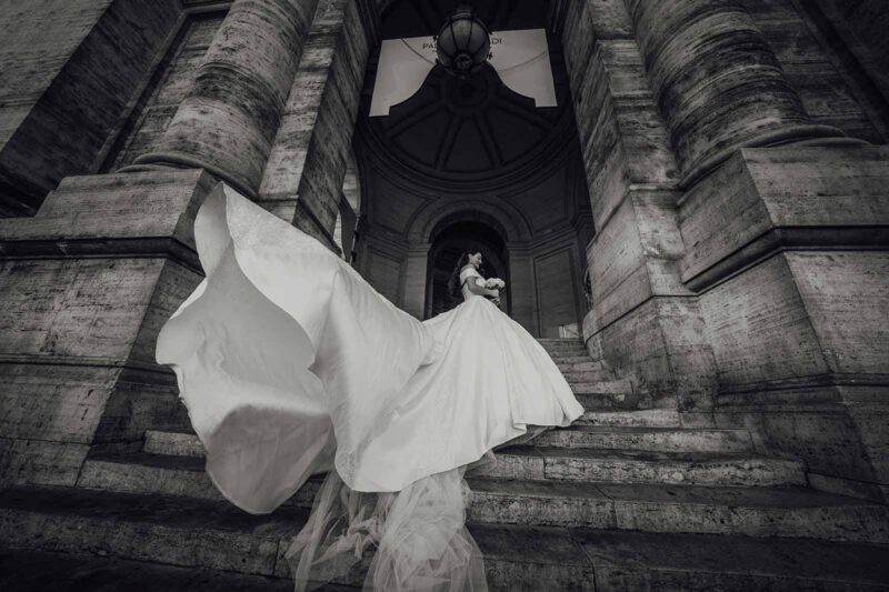 Elegant bride in a flowing white gown holding a bouquet at a historic stone venue, showcasing luxury wedding fashion and architecture.