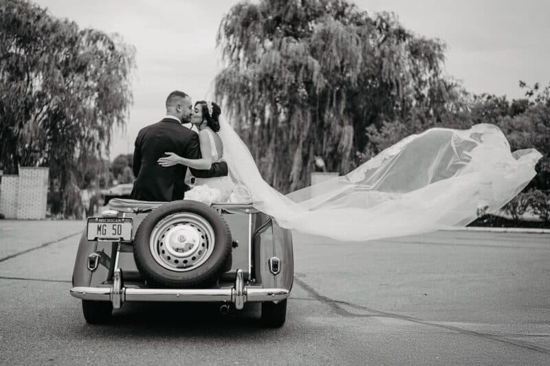 Elegant wedding couple sharing a kiss in a vintage car, celebrating a luxurious wedding day outdoors at a high-end venue.