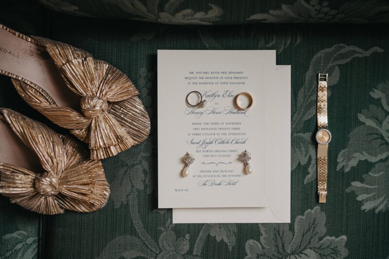 Luxurious wedding invitation with elegant rings, pearl earrings, and stylish accessories on a dark textured surface.
