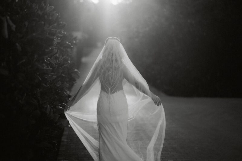 Elegant bride walking outdoors in a flowing wedding gown with veil, captured in black and white at sunset.