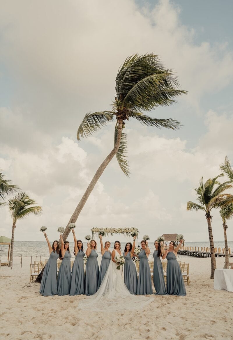 Elegant beach wedding with bridesmaids in matching slate gowns, under swaying palm trees on sandy shores.