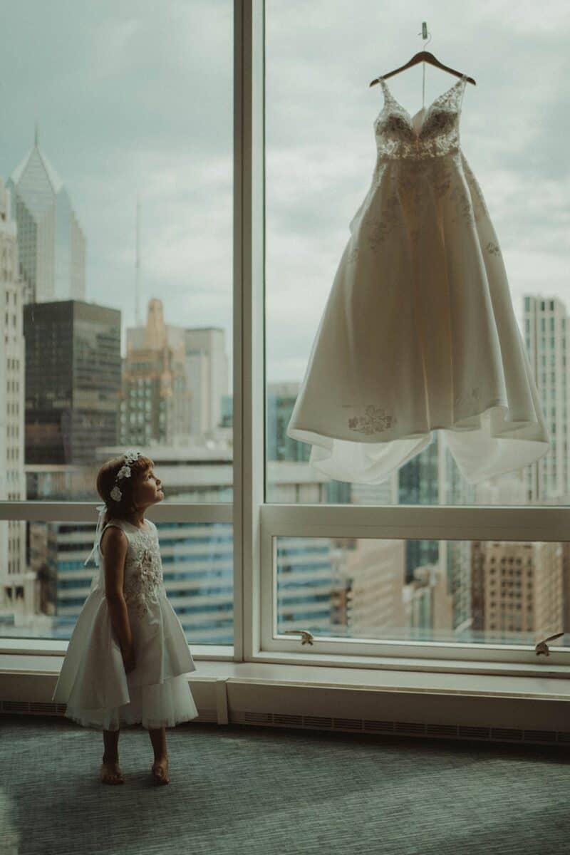 Luxury wedding dress hanging in high-rise city hotel room with young girl in floral dress looking at it, urban skyline in background.