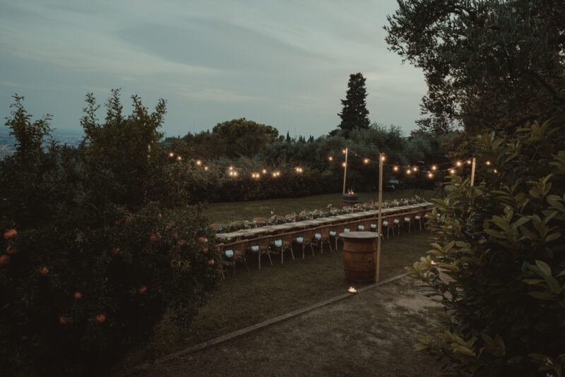 Elegant outdoor wedding reception setup with string lights and a long banquet table amidst lush greenery at dusk.
