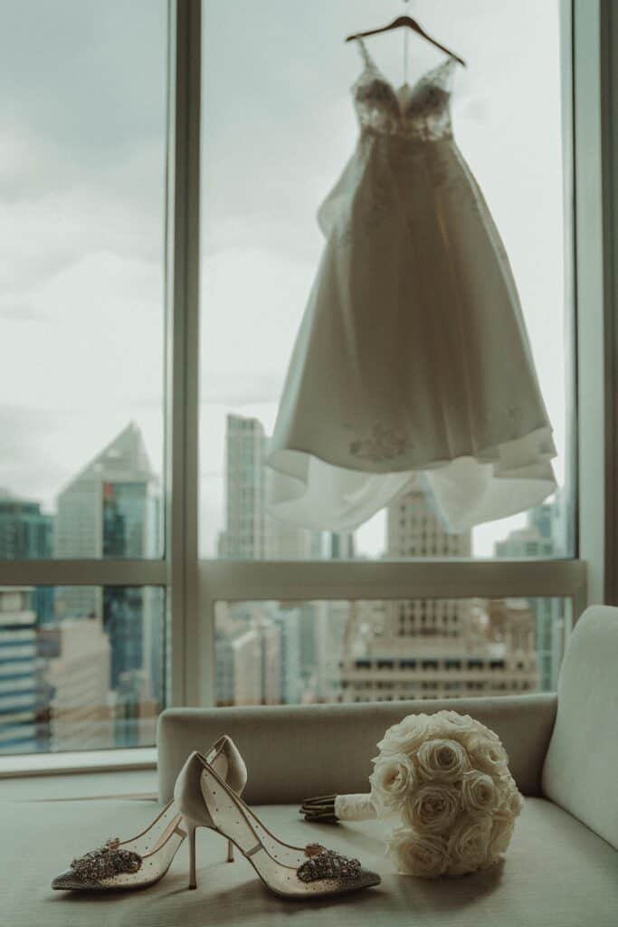 Bride's wedding shoes, bouquet of white roses, and wedding dress on hanger against city skyline window.