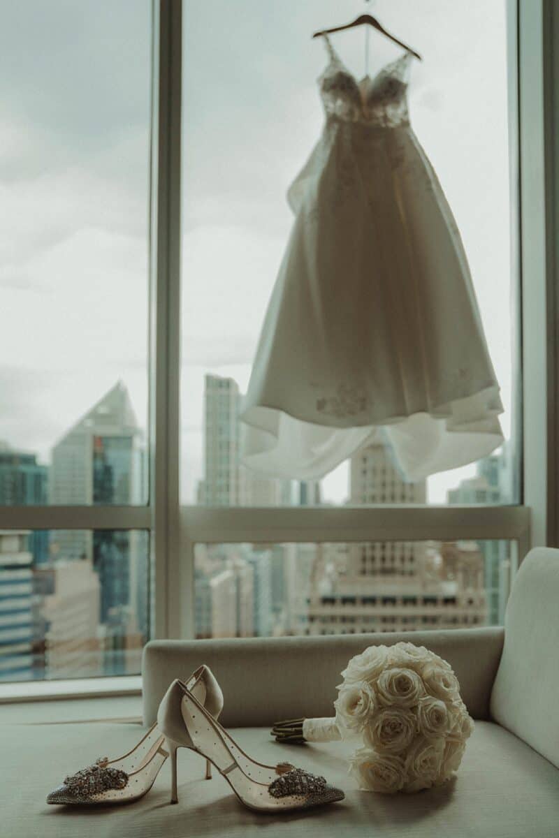 Bride's wedding shoes, bouquet of white roses, and wedding dress on hanger against city skyline window.
