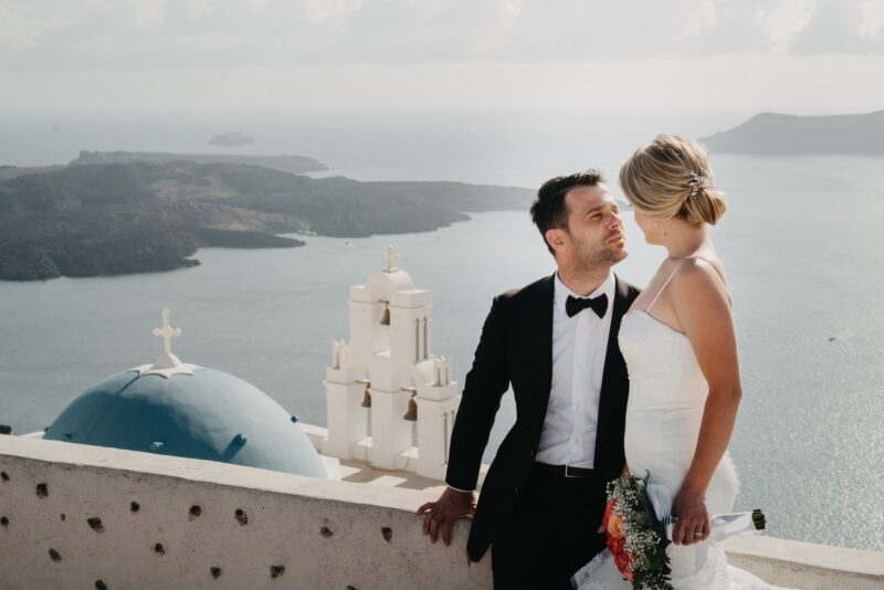 Elegant couple in wedding attire overlooking Santorini's iconic blue-domed church, capturing a romantic moment with the stunning Aegean Sea in the background.