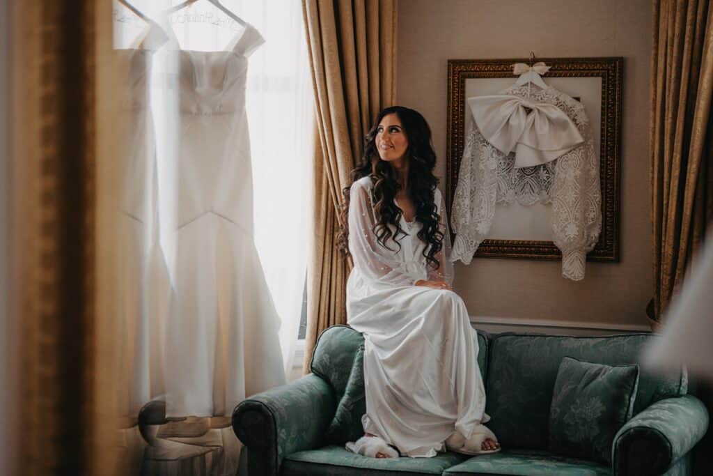 Elegant bride in a white silk gown sitting on a vintage sofa near her wedding dress and lace accessories in a luxurious hotel room.