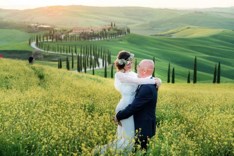 Florence countryside wedding couple embracing among yellow flowers with rolling hills and cypress trees in the background.