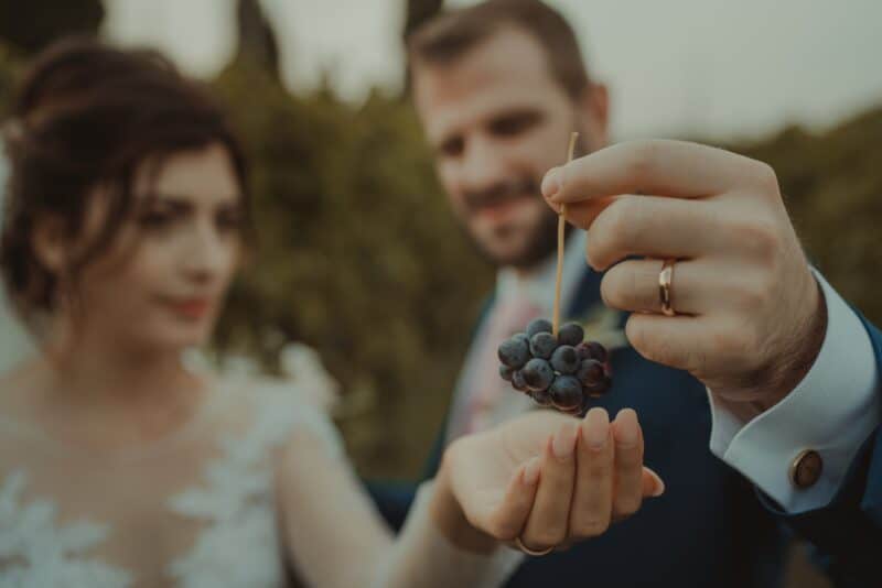 Groom holding a bunch of grapes at a wedding, high-end outdoor venue, romantic wedding scene with bride and groom, luxury wedding photography, elegant wedding celebration in nature, stylish wedding couple, outdoor vineyard wedding setting, sophisticated wedding ambiance.