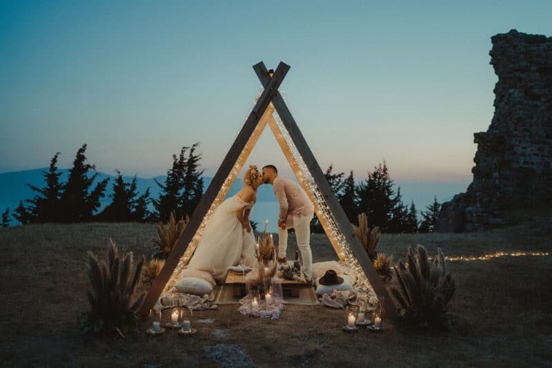 Elegant outdoor wedding ceremony with a rustic wooden tipi, romantic string lights, and scenic mountain backdrop at sunset.
