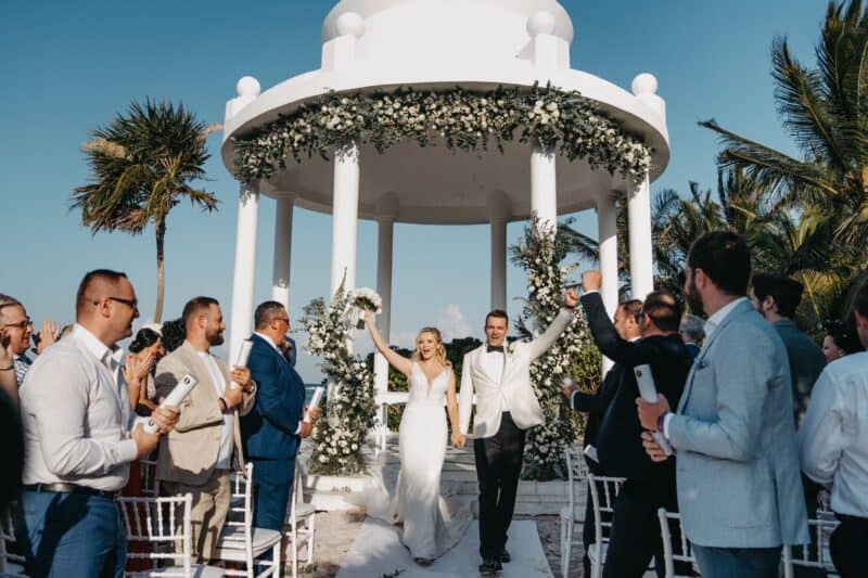 Elegant wedding ceremony at a luxurious seaside venue with a white floral gazebo, capturing a joyful newlywed couple and guests celebrating.