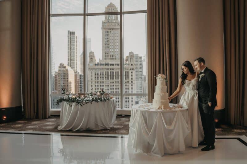 Elegant wedding couple cutting a high-end tiered wedding cake in a luxurious city skyline ballroom.