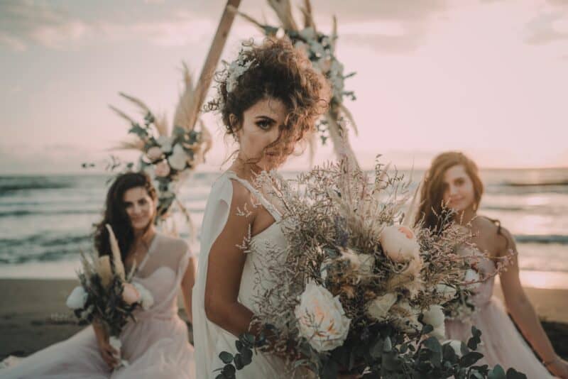 Elegant bride holding a lush bouquet at sunset on the beach with two bridesmaids, showcasing a luxurious wedding setting and high-end floral arrangements.
