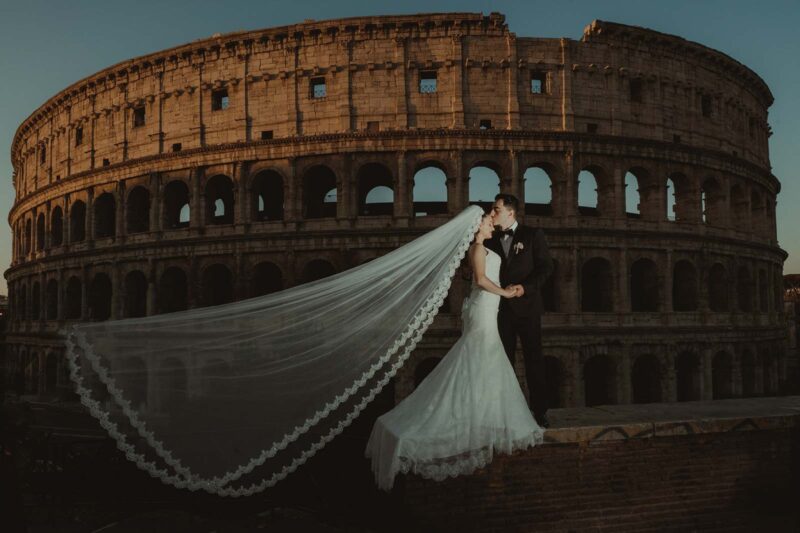 Romantic wedding couple kissing in front of the Colosseum at sunset for a luxurious high-end wedding photoshoot.