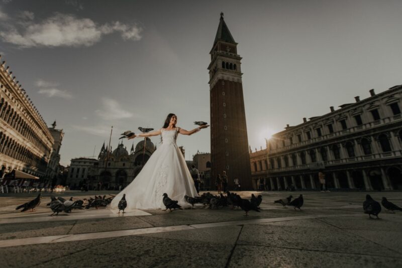 Elegant bride in a white wedding gown surrounded by pigeons at Piazza San Marco, Venice, during sunset.