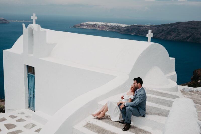 Stunning couple sitting outside a whitewashed church with blue door overlooking the Aegean Sea in Santorini, Greece. Ideal for luxury destination weddings.