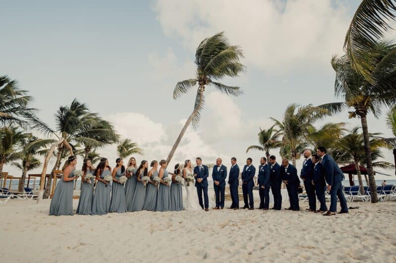 Luxurious beach wedding with elegantly dressed bridal party under palm trees on a sunny day.