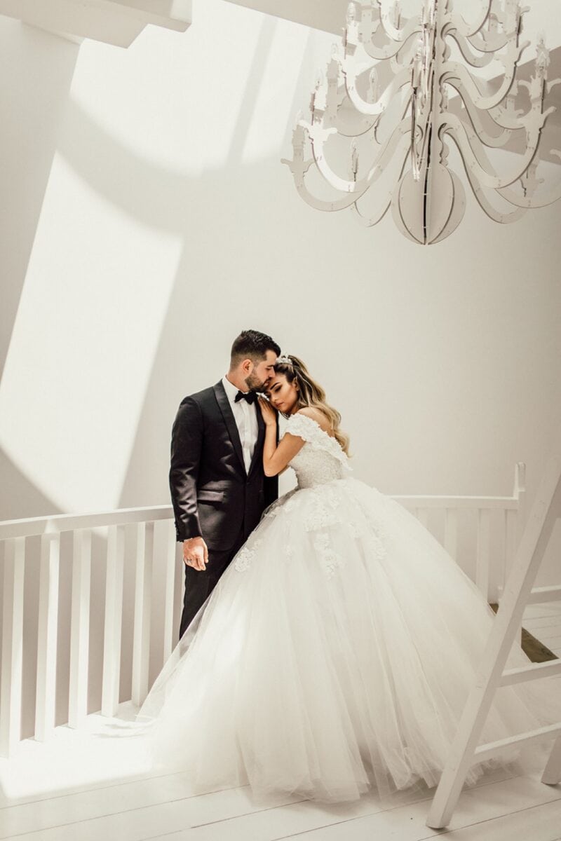 Elegant wedding portrait in a luxurious high-end venue featuring a bride in a voluminous gown and groom in a tuxedo under a grand chandelier.