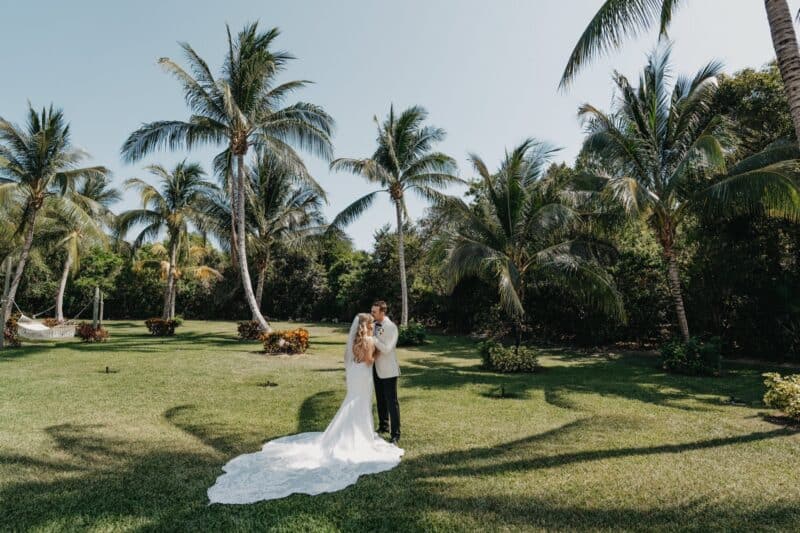 Elegant wedding couple embracing under palm trees at luxurious outdoor tropical venue.