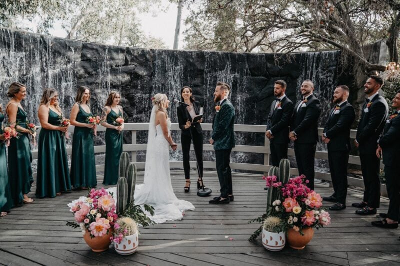 Elegant outdoor wedding ceremony at a luxury venue with cascading waterfall backdrop, featuring stylish formal attire, vibrant floral arrangements, and a sophisticated ambiance.