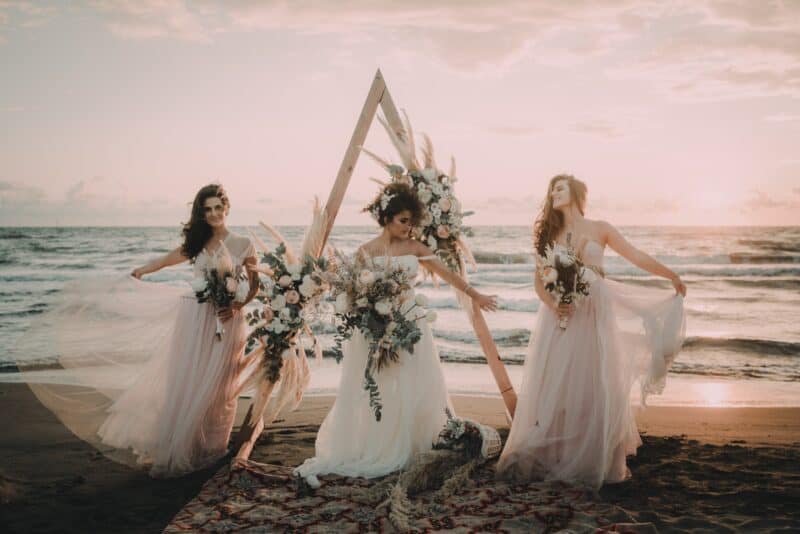 Elegant bridal party on the beach during sunset with floral arch and flowing dresses.