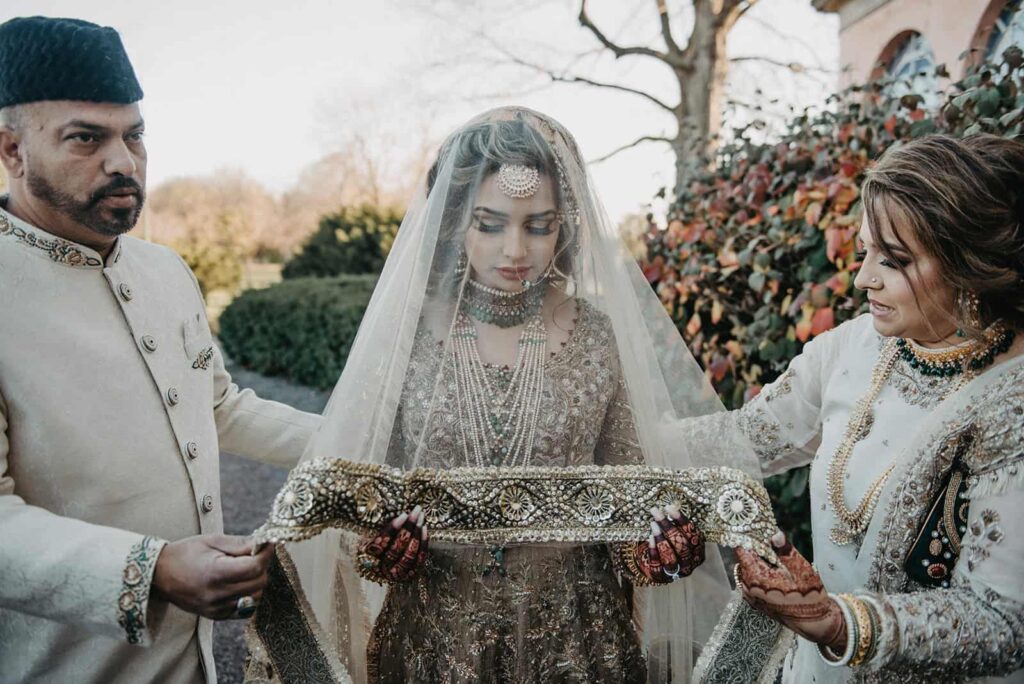 Elegant Indian bride adorned with jewelry and traditional attire, surrounded by her family during wedding ritual outdoors.