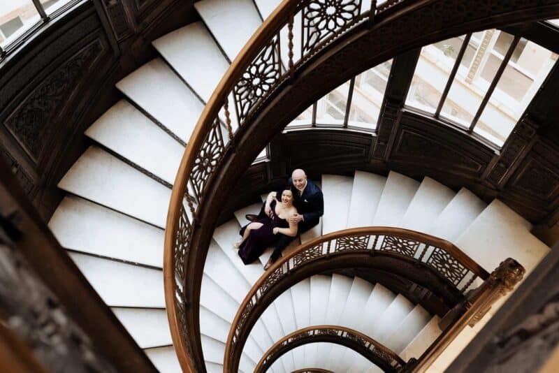 Elegant couple on grand spiral staircase at luxury historic venue during wedding celebration.