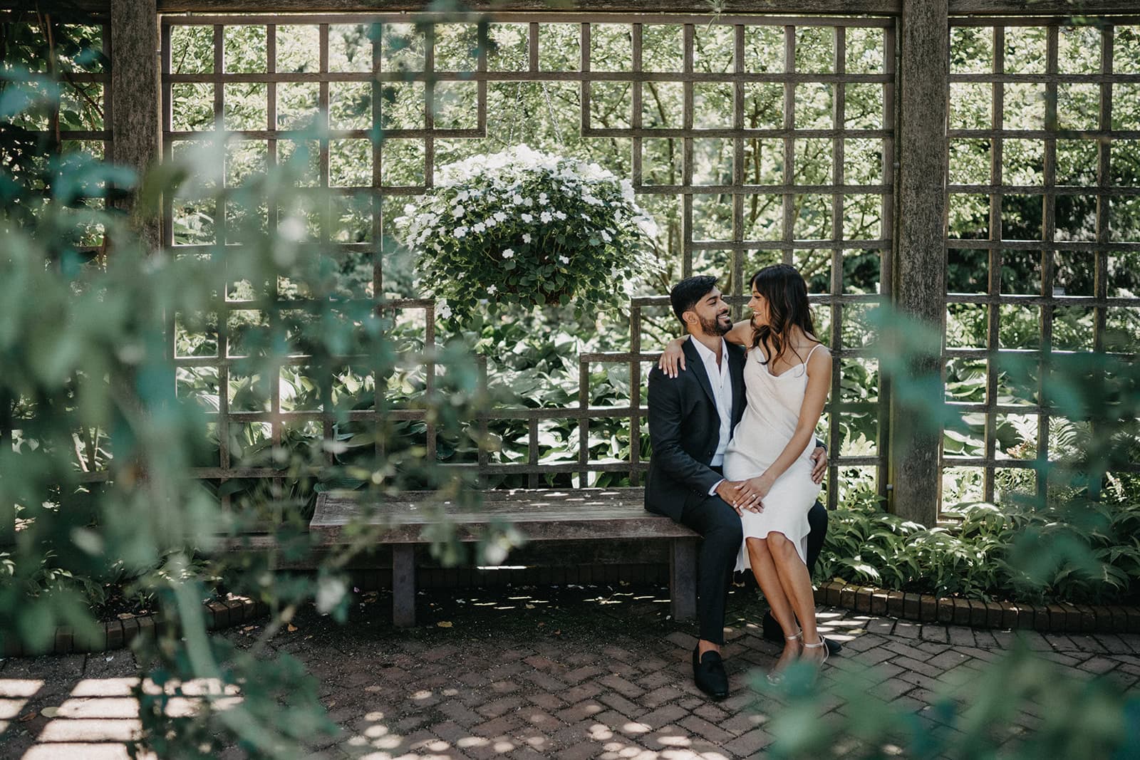 Elegant couple sitting on a wooden bench outdoors amidst lush greenery, showcasing luxury wedding photography at a high-end garden venue.