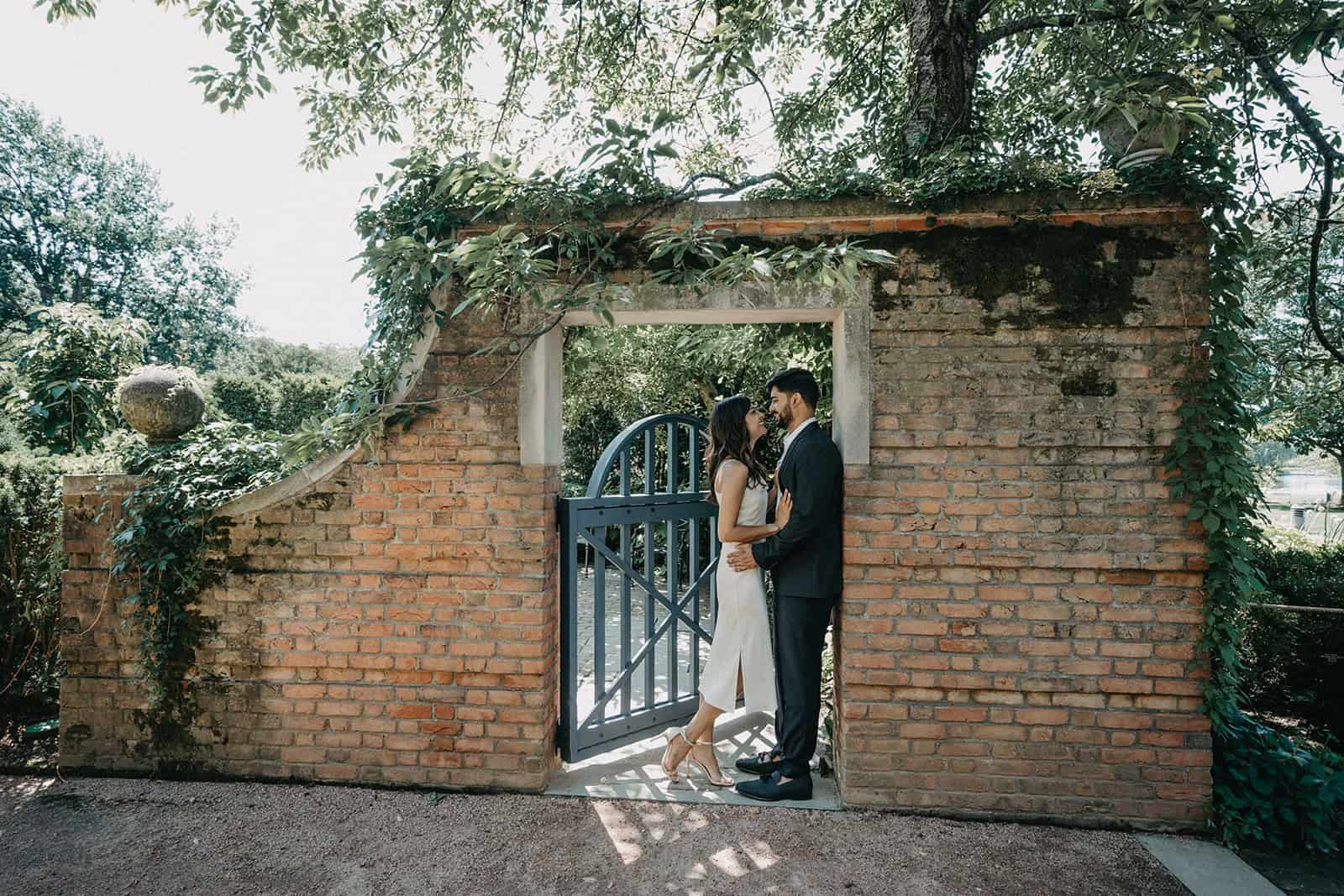 Romantic couple in elegant wedding attire standing at a rustic brick gate amidst lush greenery, perfect for luxury wedding venues.