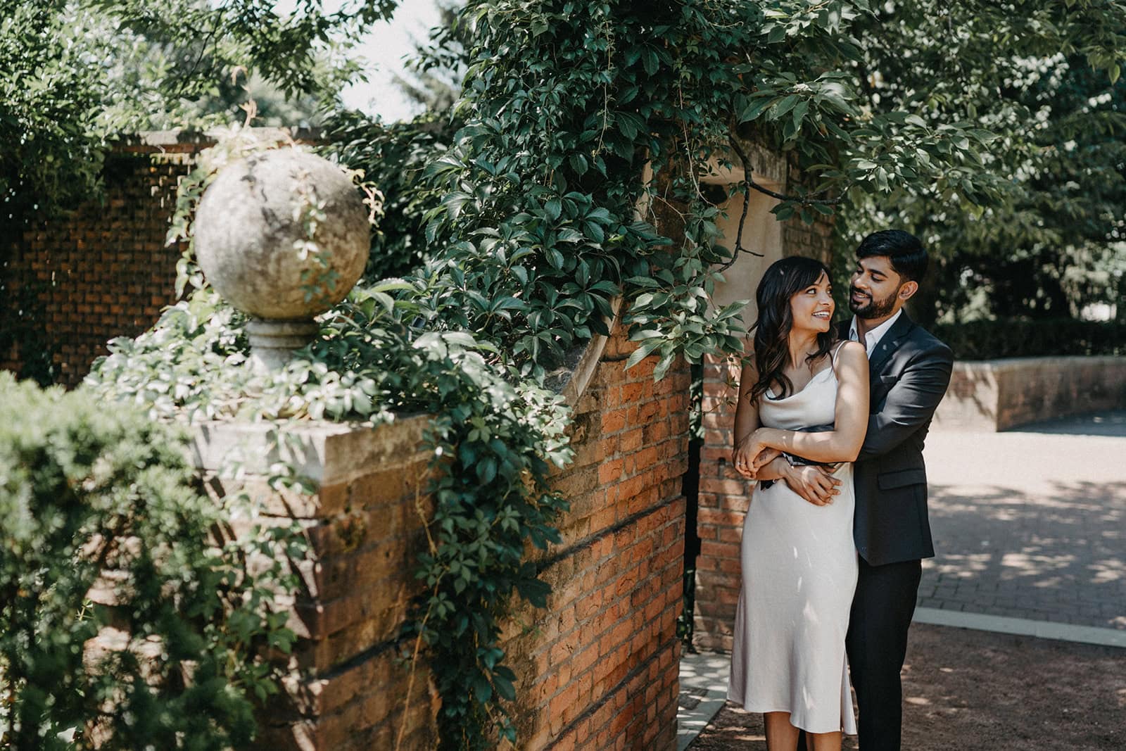 Elegant couple in wedding attire sharing a moment outdoors against a lush greenery and brick wall backdrop.