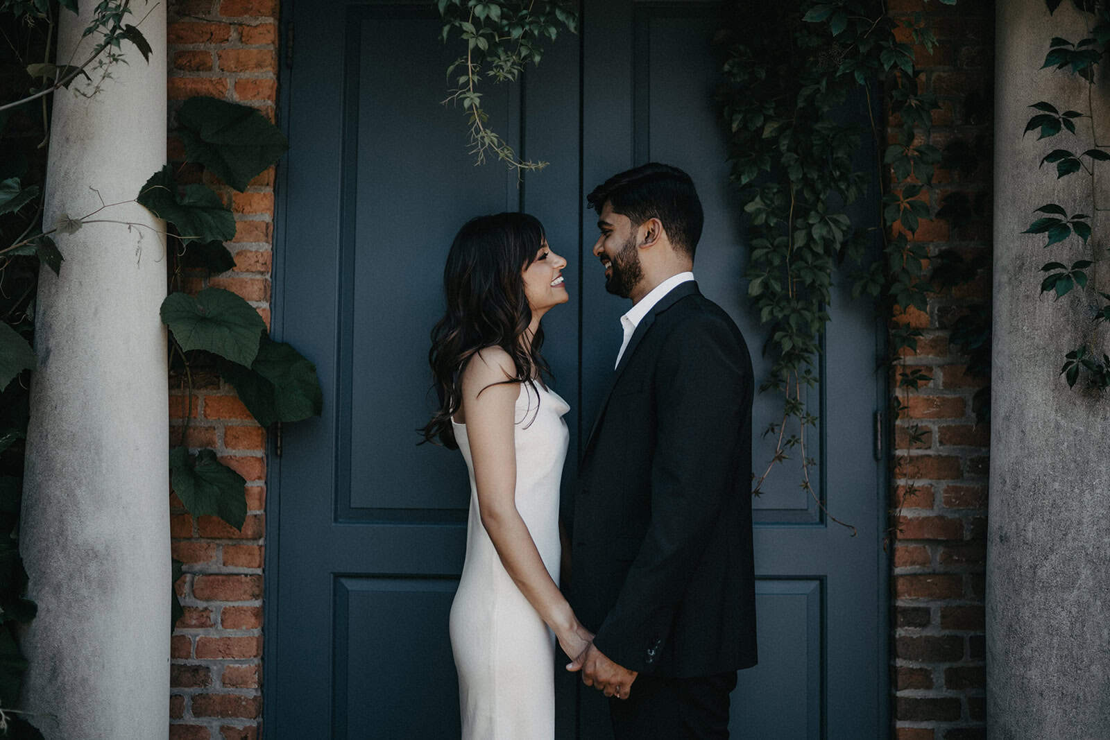 Elegant couple in wedding attire standing face-to-face in front of a stylish blue door with lush greenery, perfect for luxury wedding photography.