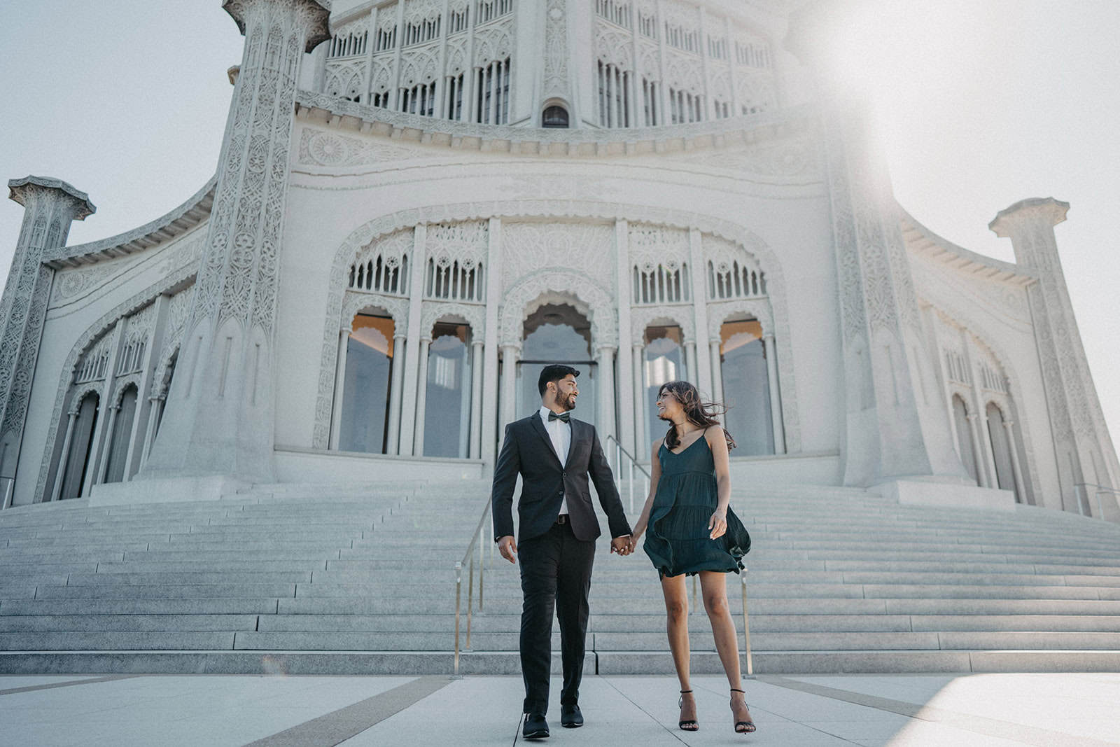 Elegant couple in formal wear holding hands on grand staircase at luxurious wedding venue.
