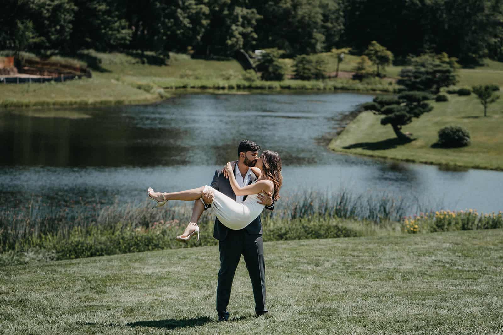 Romantic couple in wedding attire by a serene lake at a luxury outdoor wedding venue.