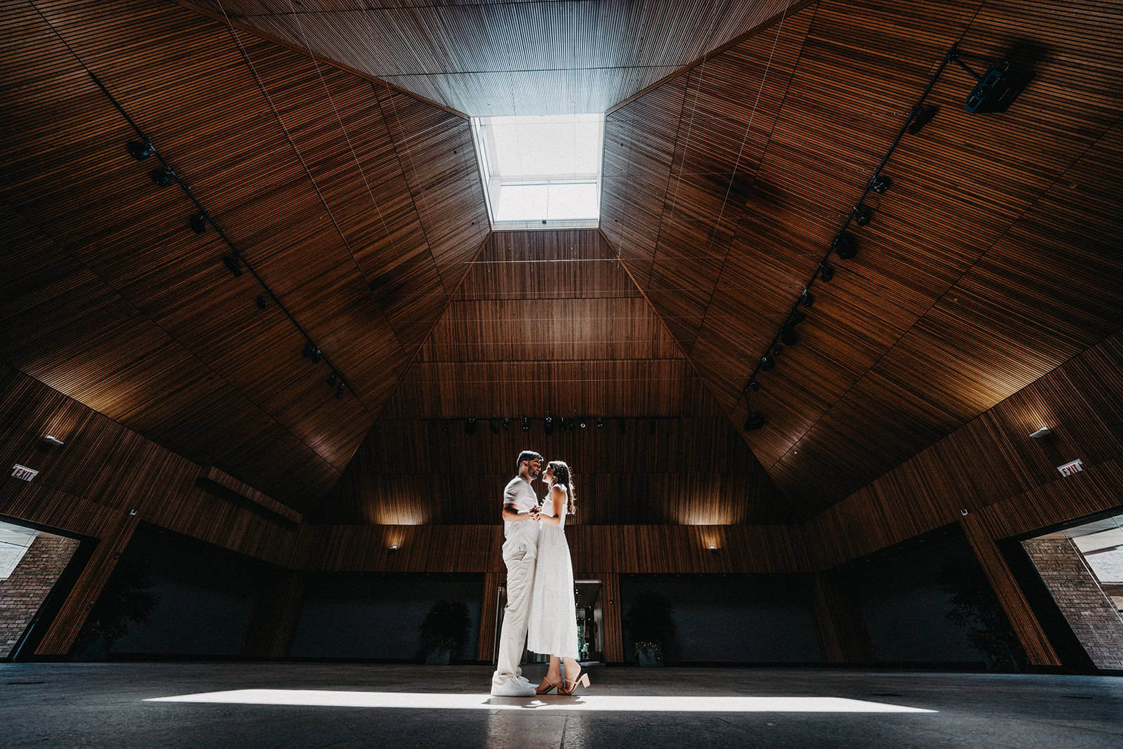 Elegant couple sharing a moment in a modern, wood-paneled wedding venue with natural lighting from a skylight.