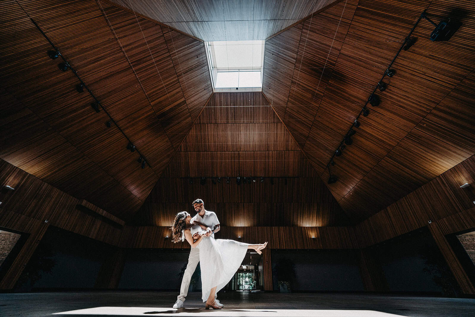 Elegant couple dancing inside a luxurious, modern venue with a stunning wooden vaulted ceiling and natural skylight.