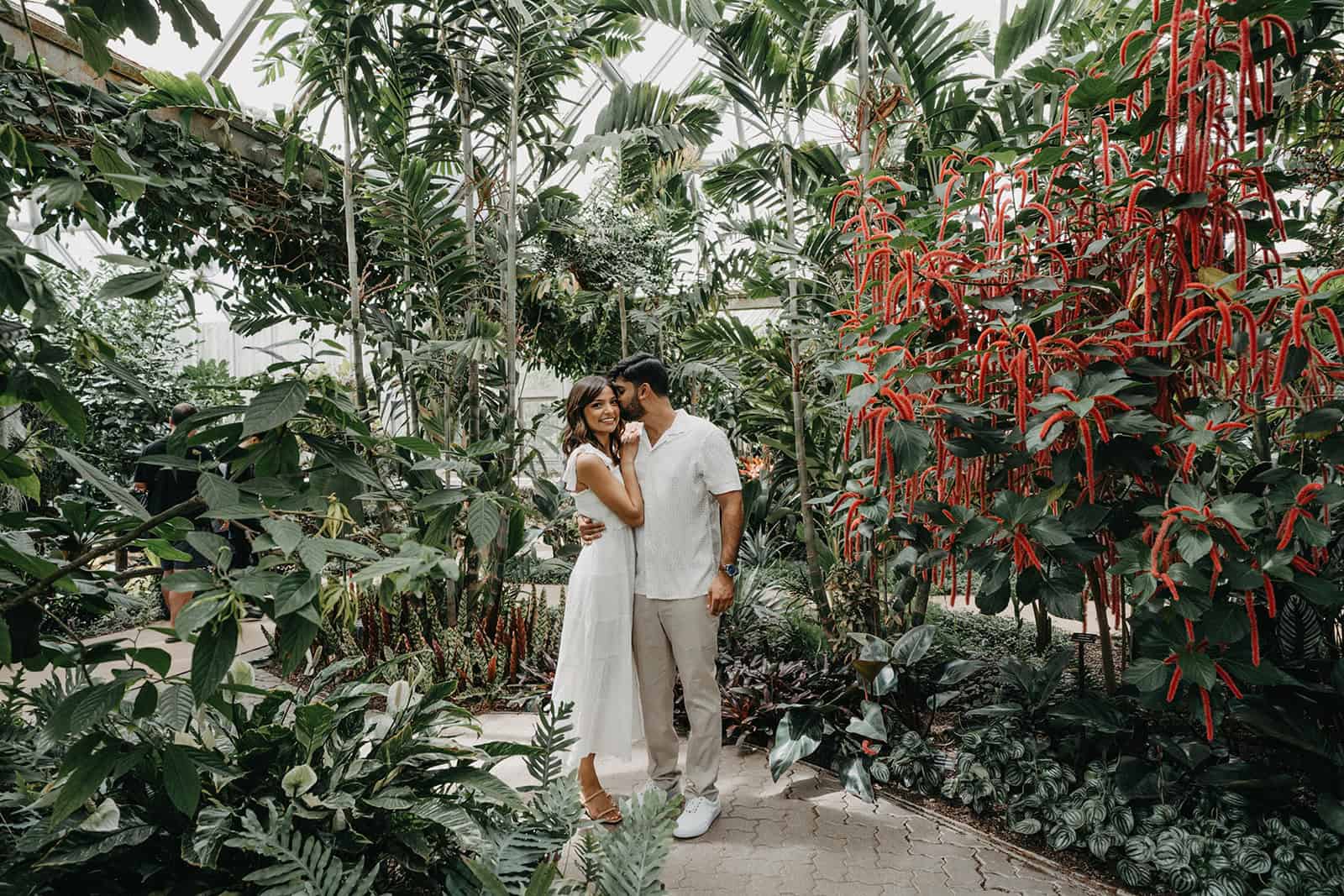 Lush tropical greenhouse wedding backdrop with couple in elegant white attire, surrounded by exotic plants and vibrant flowers.